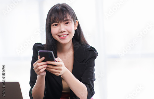 Confident young Asian woman using smartphone while working on laptop computer in home office. Remote working, freelancer, small business concept