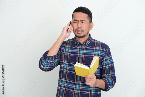Adult Asian man try to remember something while holding a book and pen photo