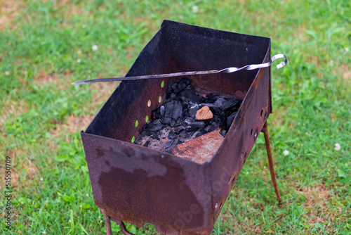 rusted metal grill with metal legs in green grass with a silver skewer across it. Black unburnt coals remained in the grill. photo