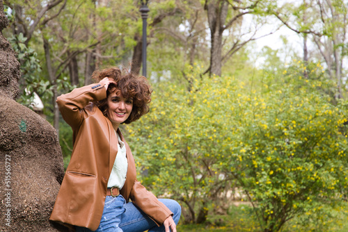 beautiful Spanish woman with curly hair is leaning on a tree trunk in the park. The woman is happy and enjoying her holiday in the city. Concept happiness, fashion and beauty. © @skuder_photographer