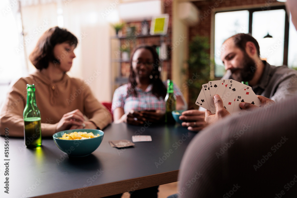 Joyful multicultural group of friends sitting at table while playing card games together. Selective focus shot of man enjoying fun leisure activity with people while having beer and snacks.