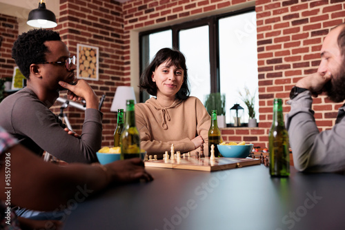 Cheerful woman sitting with friends at home in living room while playing chess. Happy diverse people enjoying strategic boardgames together while relaxing and consuming snacks and beverages.