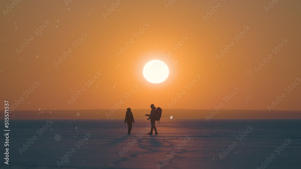 Fototapeta premium The two travelers walking through the snow field on sunset background