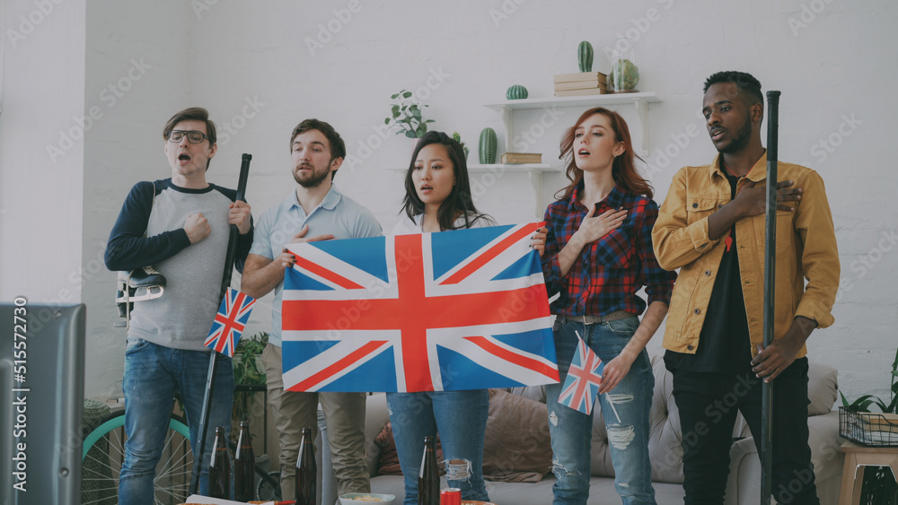 Multi ethnic group of friends sport fans listening and singing British national anthem before watching sports championship on TV together at home indoors - obrazy, fototapety, plakaty 