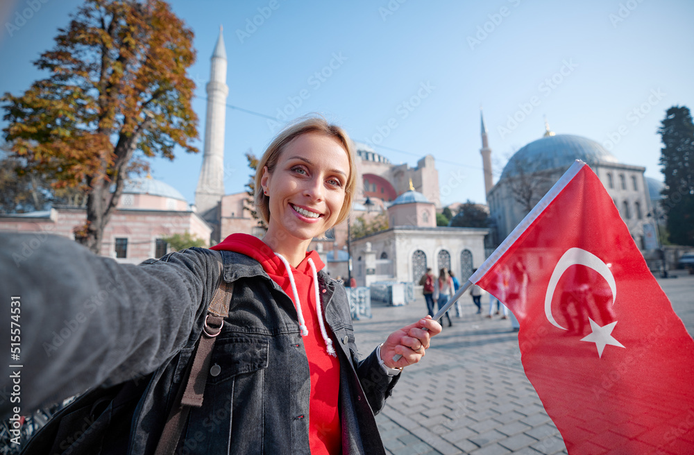Enjoying vacation in Istanbul. Young traveling woman with national turkish flag taking selfie on view of Hagia Sophia Temple. - obrazy, fototapety, plakaty 