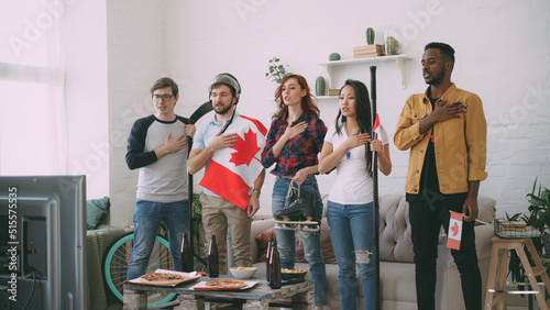 Multi ethnic group of friends sport fans listening and singing national Canadian anthem before watching sports championship on TV together at home indoors photo