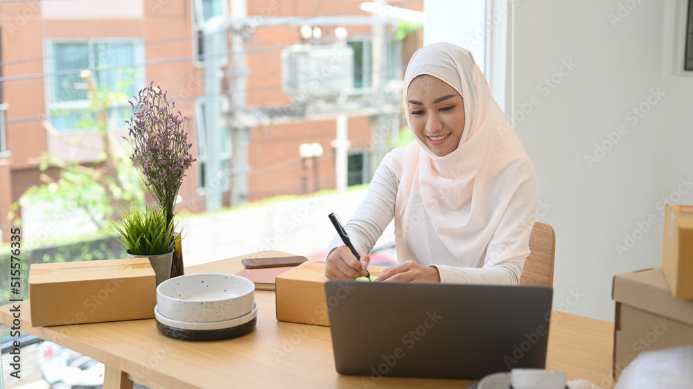 Muslim Woman in hijab checking product purchase order on laptop and preparing parcel boxes of product for delivery to customers Muslim Woman in hijab checking product purchase order on laptop and preparing parcel boxes of product for delivery to customers
