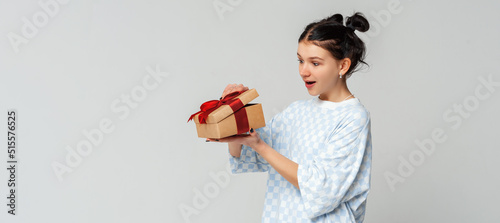 Portrait of a happy smiling young dark-haired girl opening a gift box, standing in trendy t shirt over light grey background