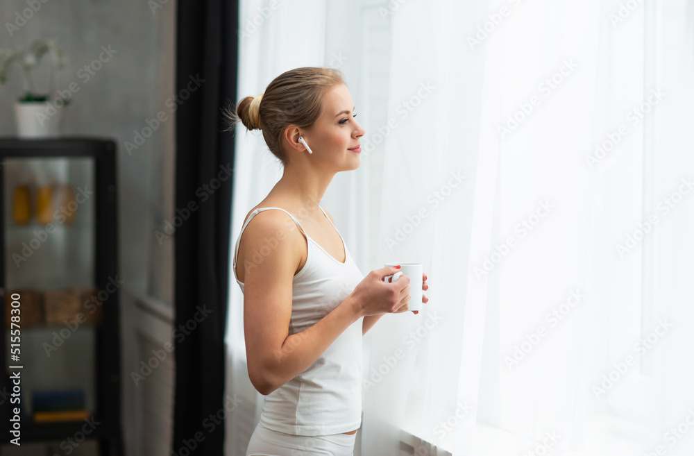 Young woman in white underwear is standing in front of the window and looking into it. Girl at home in the living room. Young woman in white underwear is standing in front of the window and looking into it. Girl at home in the living room.