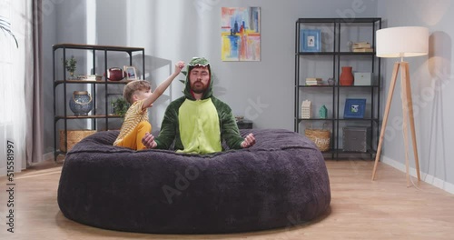 Caucasian handsome man father in green dragon costume sitting on roung soft couch and meditating, then laughing and playing with small cute son. Litle boy having fun with dad at home. photo