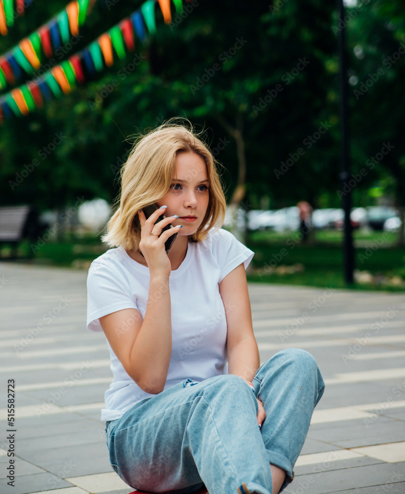 A young beautiful teen girl is sitting on a skateboard on a summer day in the park. A girl is talking on a mobile phone in the open air. Chat with friends. A young beautiful teen girl is sitting on a skateboard on a summer day in the park. A girl is talking on a mobile phone in the open air. Chat with friends.