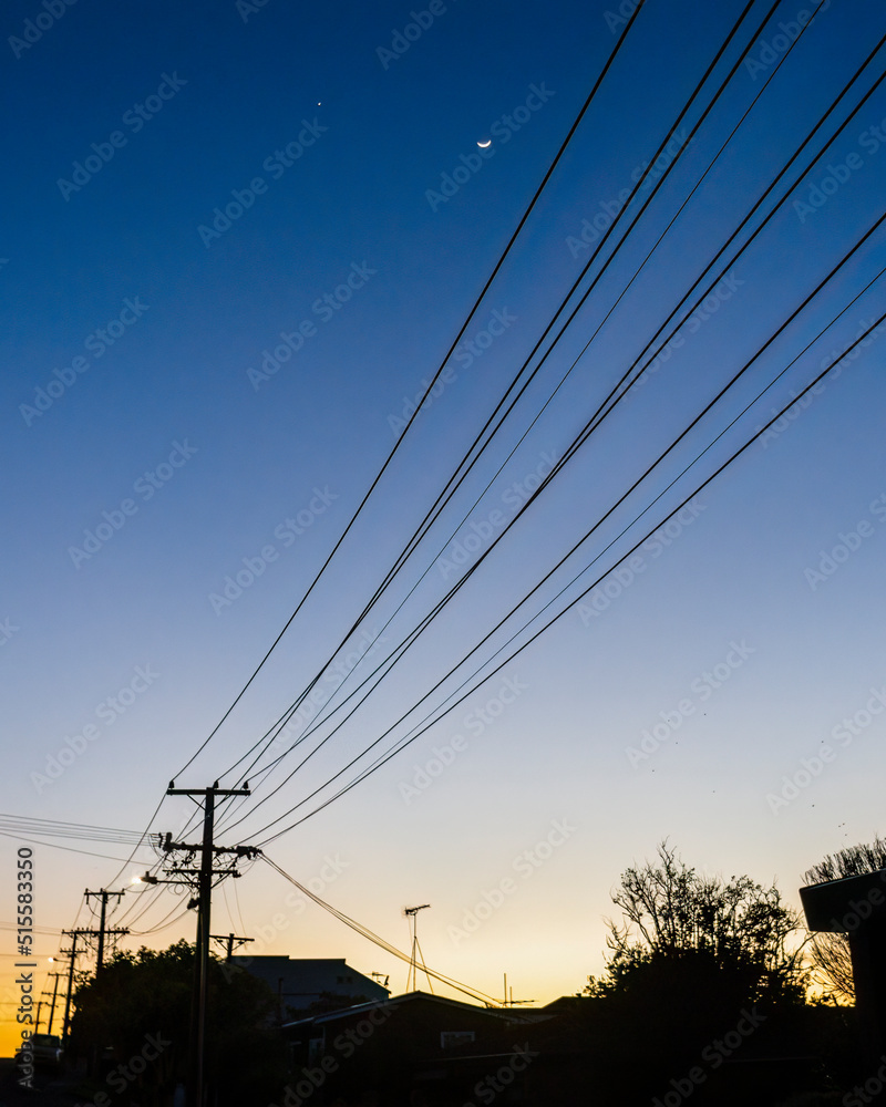 Waxing crescent moon over silhouette powerlines at dawn. Auckland. Vertical format.