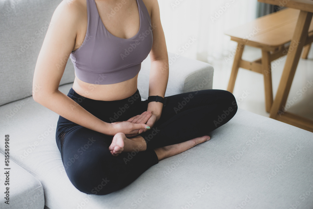 Fototapeta premium Woman wearing black sport wear sitting on sofa practicing yoga and meditating in living room. She preparing material for practice class. Calmness and relaxation. Female happiness and yoga concept.