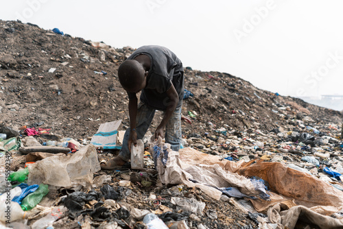 Unrecognizable adolescent in a worn black T-shirt and tattered trousers looking for items among the trash spilled on a big landfill in West Africa