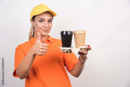 Blonde female courier holding two cups of coffees and making thumbs up sign on white background