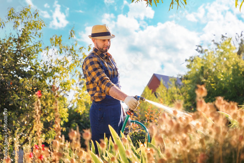 Ecofarm. A bearded, handsome young gardener in uniform and a straw hat, hosing plants and flowers. In the background there is a backyard and a garden. The concept of gardening and horticulture photo
