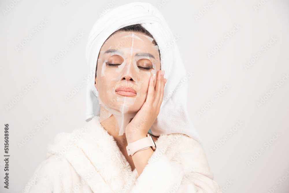 Portrait of young lady in bathrobe and towel on head while touching her face with facemask Portrait of young lady in bathrobe and towel on head while touching her face with facemask
