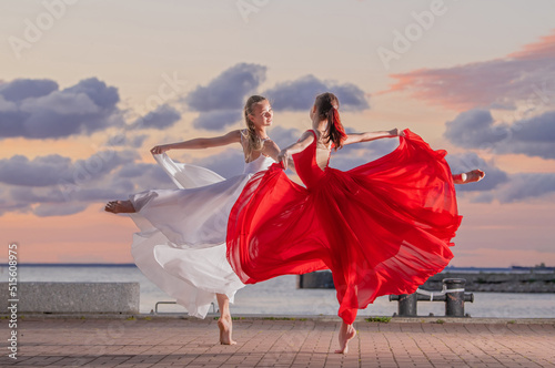 Two ballerinas in a white and red flying skirt and leotard dancing in a duet on the embankment of the ocean or sea against the backdrop of the sunset sky