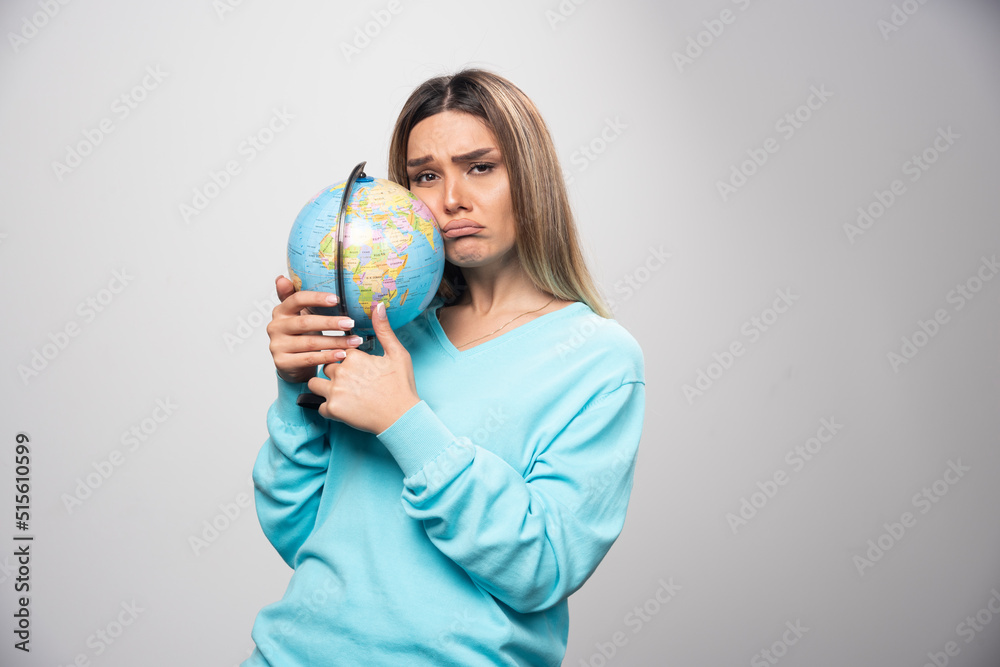 Blonde girl in blue sweatshirt holds a globe and looks uncertain and confused Blonde girl in blue sweatshirt holds a globe and looks uncertain and confused