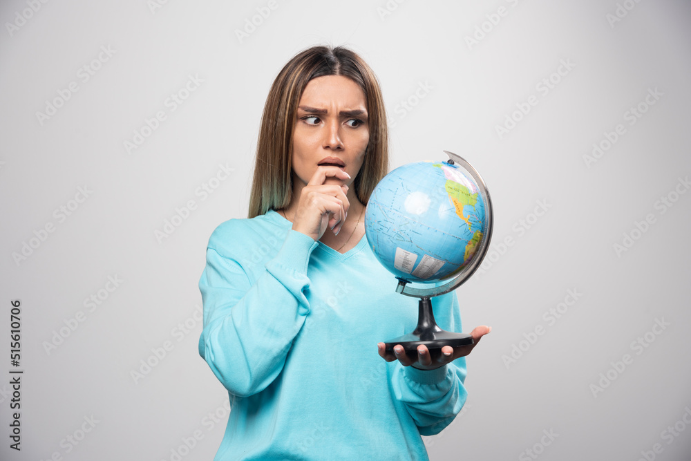 Fototapeta premium Blonde girl in blue sweatshirt holding a globe, thinking carefully and trying to remember