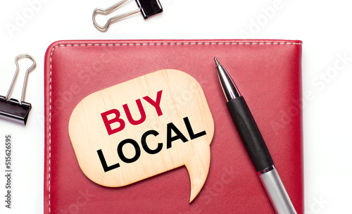 On a light background there are black paper clips, a pen, a burgundy notepad a wooden board with the text BUY LOCAL