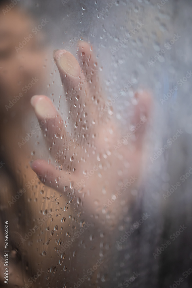 Female hand and unrecognizable silhouette behind wet glass in shower