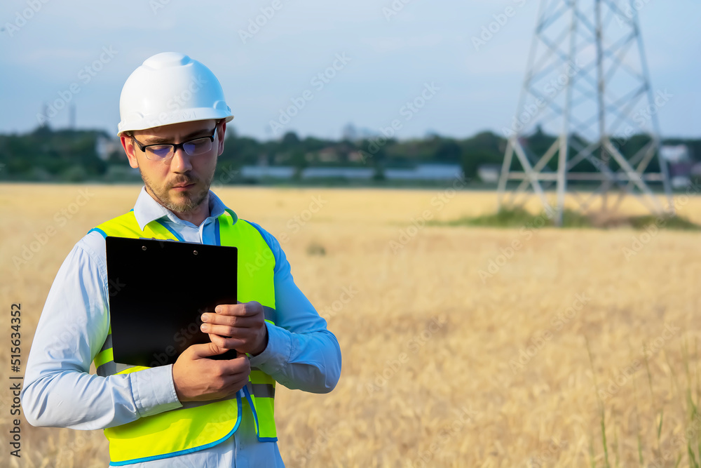 Fototapeta premium engineer in a helmet, glasses and a yellow vest, hand on a helmet, holding a notebook, a folder, making notes in a notebook a caucasian white engineer