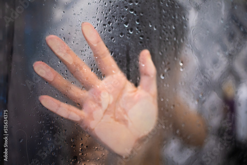 Female hand and unrecognizable silhouette behind wet glass in shower