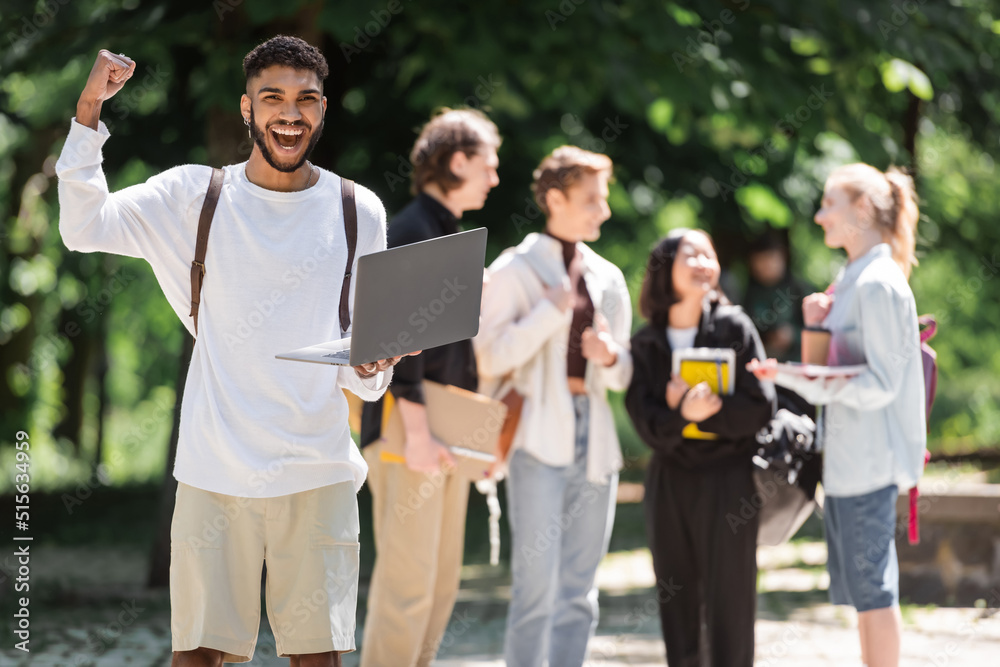 Excited african american student holding laptop near blurred friends in park.