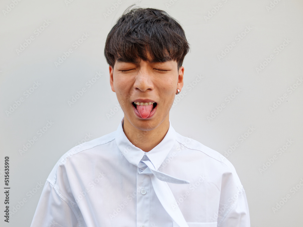 Portrait of Chinese young man with black hair in white shirt posing against white background, making face eyes closed with tongue out, dislike expression, front view. Portrait of Chinese young man with black hair in white shirt posing against white background, making face eyes closed with tongue out, dislike expression, front view.