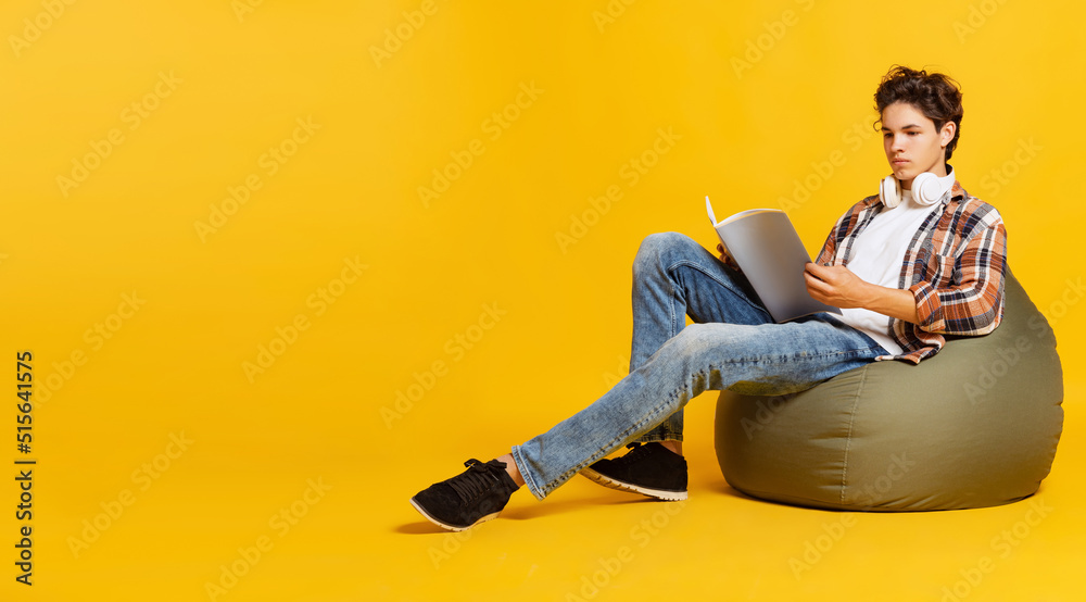 Emotive boy, student in casual style clothes sitting at chair and reading book isolated over yellow background. Back to school, university Emotive boy, student in casual style clothes sitting at chair and reading book isolated over yellow background. Back to school, university