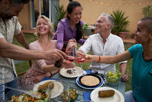 Group of friends toasting with wine having a barbecue in the backyard. Happy middle-aged people having fun at a picnic in the garden. 