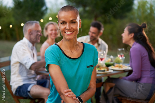 Portrait of a woman with shaved head looking at the camera. Group of friends dining outside on a terrace restaurant or backyard home. Friends having fun eating and drinking on a summer night.