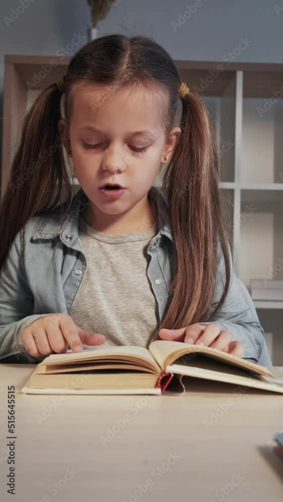 Vertical video. Child learning. Primary school training. Curious smart cute little girl reading book studying at desk at home workplace. Vertical video. Child learning. Primary school training. Curious smart cute little girl reading book studying at desk at home workplace.