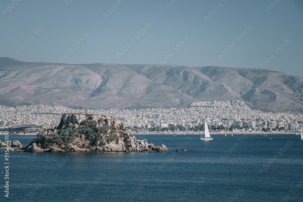 Small rock island on the mediterranean sea near the port of Athens, Greece on a sunny day and residential area and mountains in the background