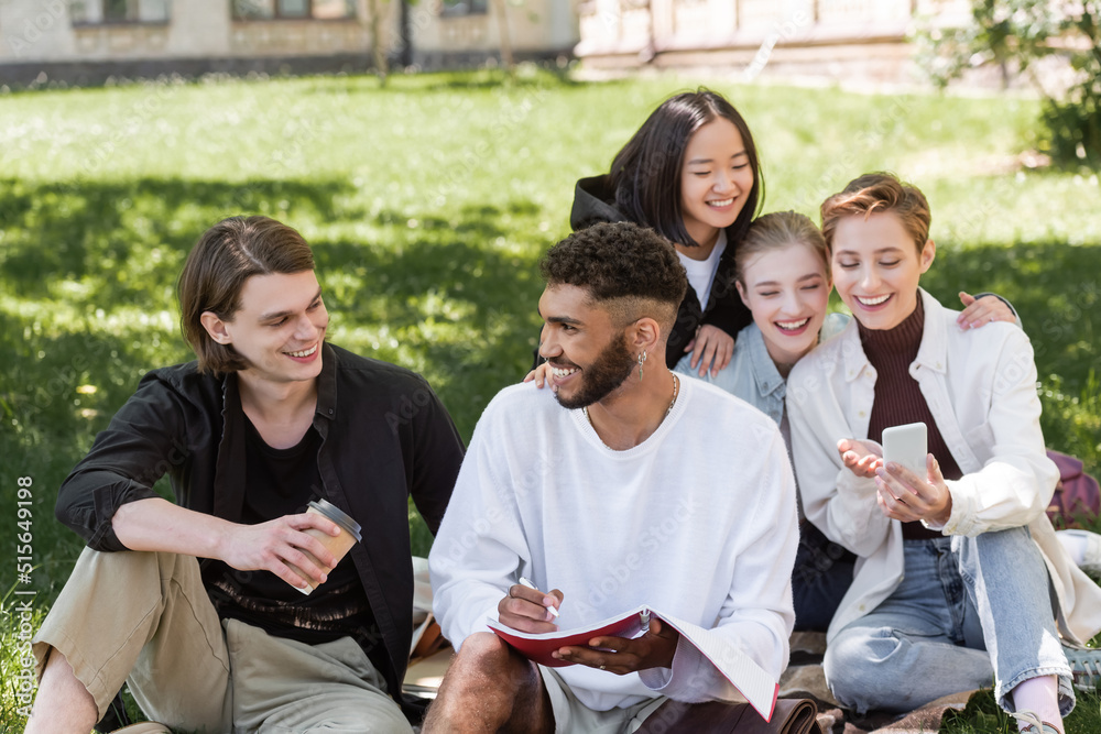 Smiling interracial students talking near blurred friends using smartphone on lawn in park.