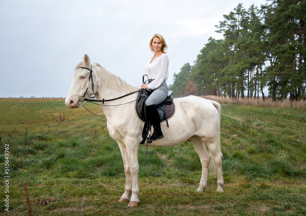 smiling blonde beautiful woman riding  white blue eyed  horse horse in green field