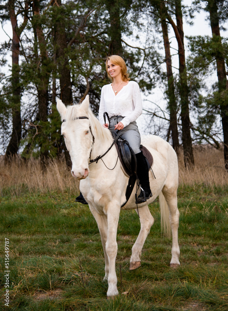 smiling blonde beautiful woman riding  white blue eyed  horse horse in green forest 