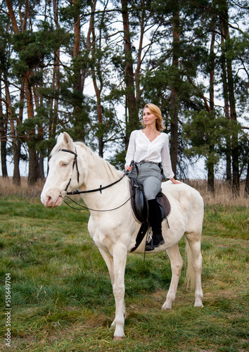 smiling blonde beautiful woman riding white blue eyed horse horse in green forest 