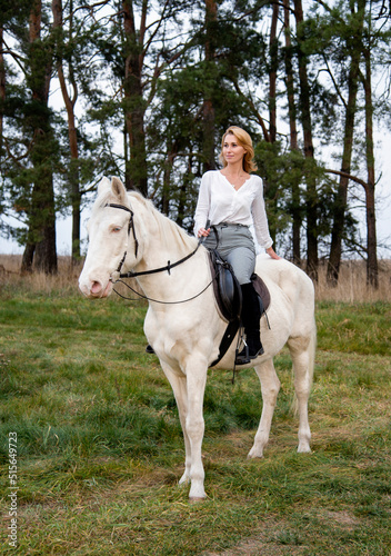 smiling blonde beautiful woman riding white blue eyed horse horse in green forest 