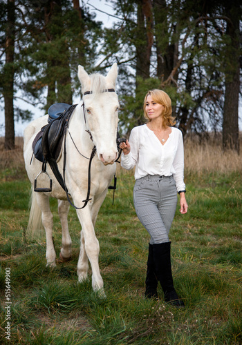 smiling blonde beautiful girl walking with white blue eyed horse horse in green forest 