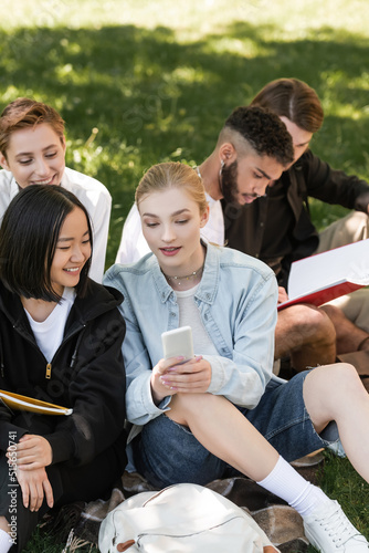 Student holding smartphone near multicultural friends on lawn in summer park.