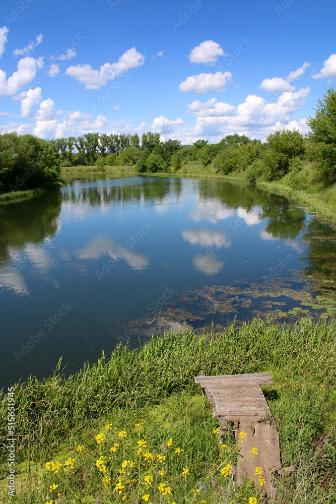 Fototapeta premium European landscape. A small calm river and a wooden fishing pier on the shore. Clouds reflect in the river.
