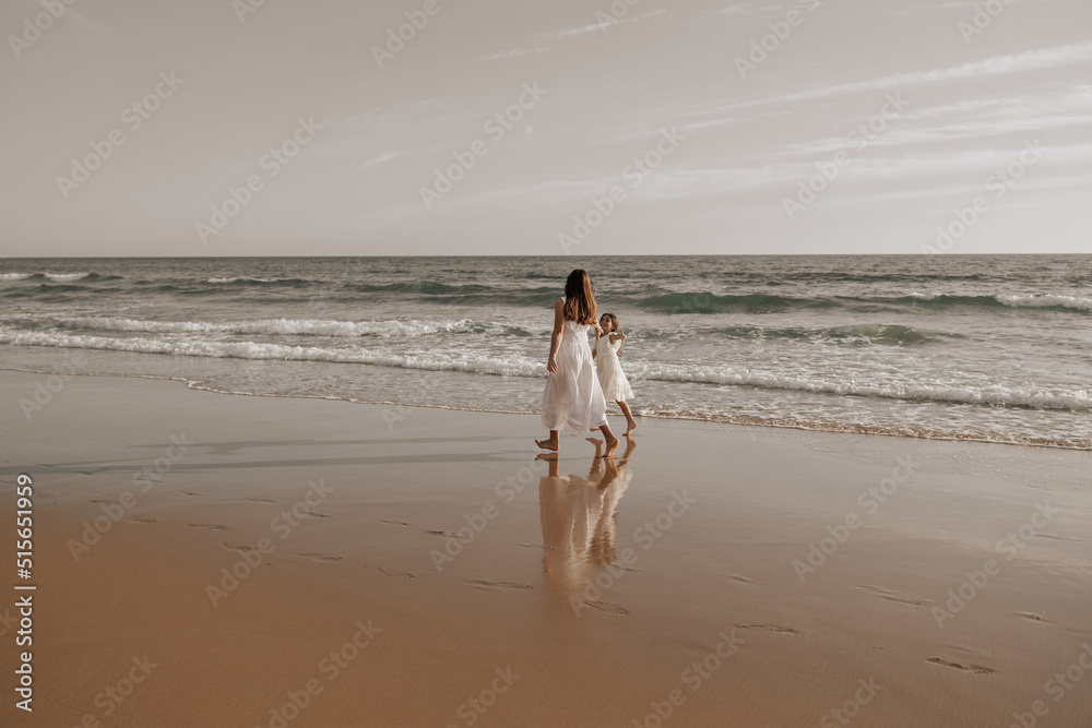 Mom and daughter in similar white dresses walking on sandy seashore Mom and daughter in similar white dresses walking on sandy seashore