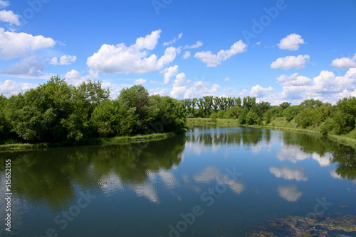European landscape. A small calm river and densely forested banks. Clouds reflect in the river.