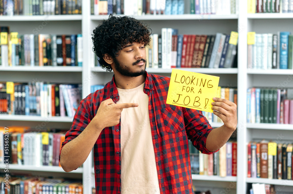 Unemployed indian guy, in casual wear, stands in the library against background of bookshelves, holds a sign in his hand that says looking for a job, looks and points finger at it, hopes to get a job Unemployed indian guy, in casual wear, stands in the library against background of bookshelves, holds a sign in his hand that says looking for a job, looks and points finger at it, hopes to get a job