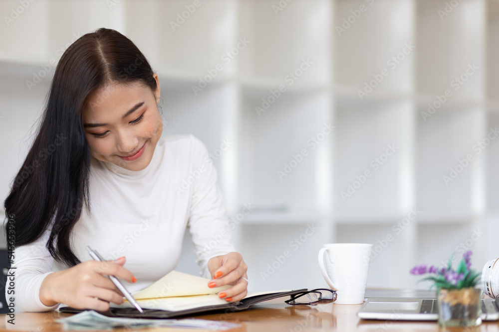 Beautiful Asian woman in the home office thinking and writing a schedule on a notepad. Beautiful Asian woman in the home office thinking and writing a schedule on a notepad.