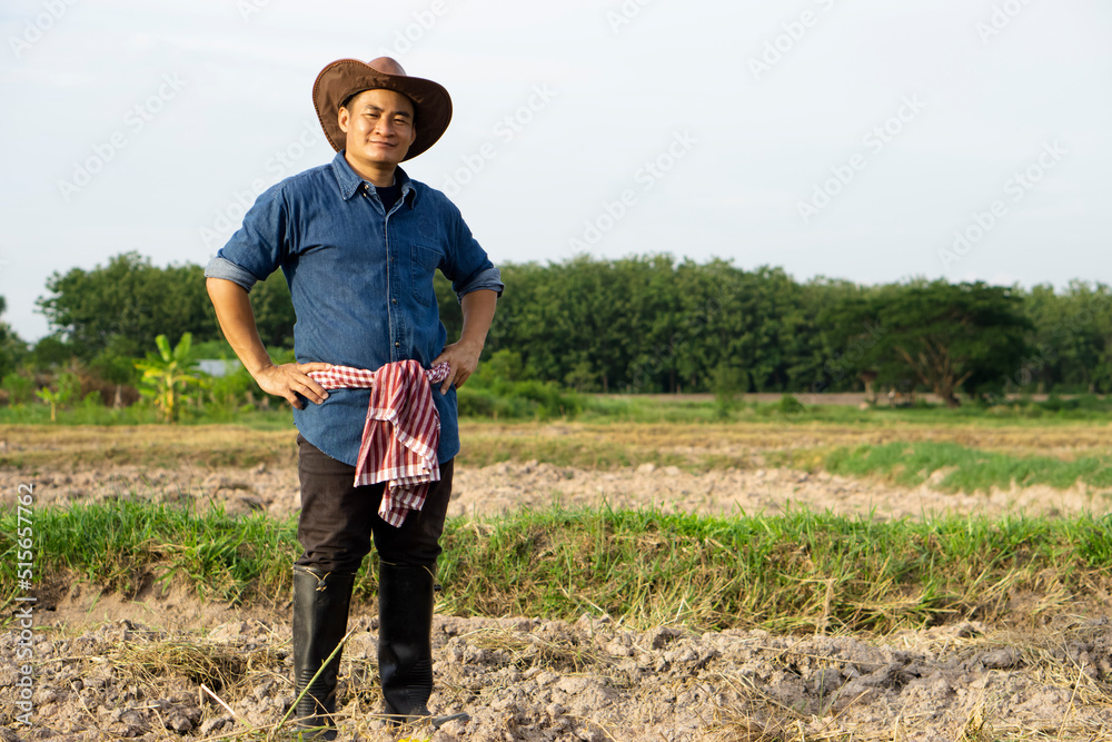 Handsome Asian man farmer is at plantation. He wears hat, blue shirt, Thai loincloth on waist, boots, put hands on hips, feels confident, looks at camera. Concept : Agriculture occupation. Full body.