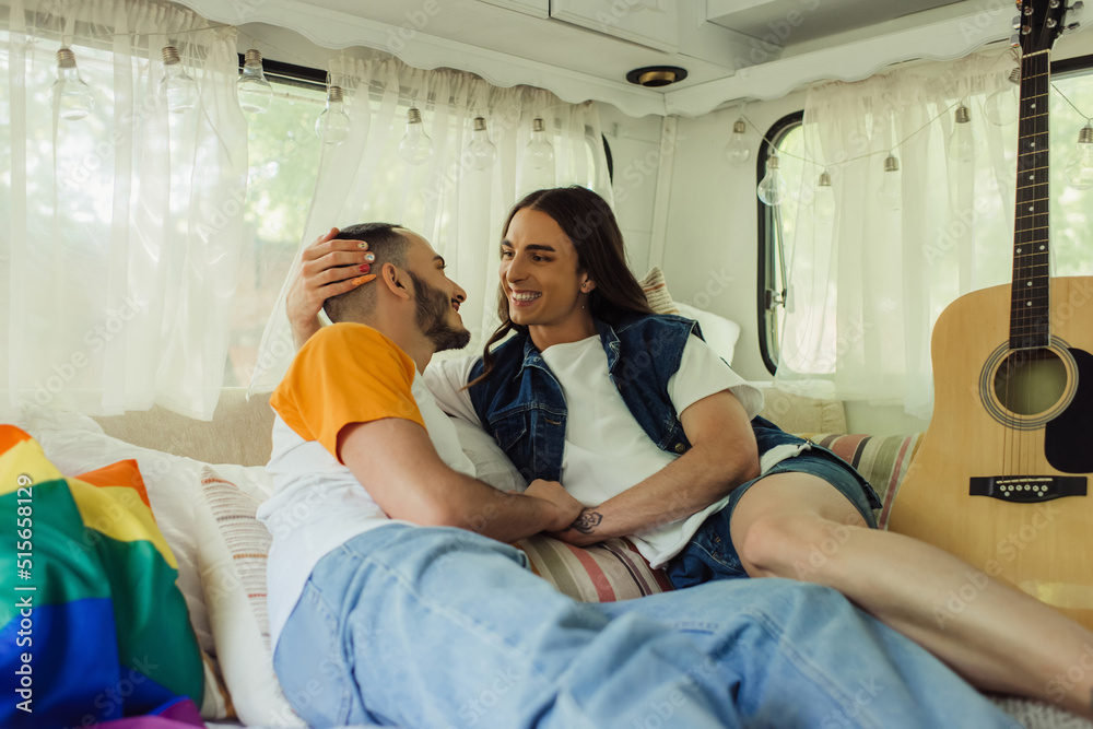 smiling gay man with tattoo looking at bearded boyfriend on bed near lgbt flag and acoustic guitar in modern van. smiling gay man with tattoo looking at bearded boyfriend on bed near lgbt flag and acoustic guitar in modern van.