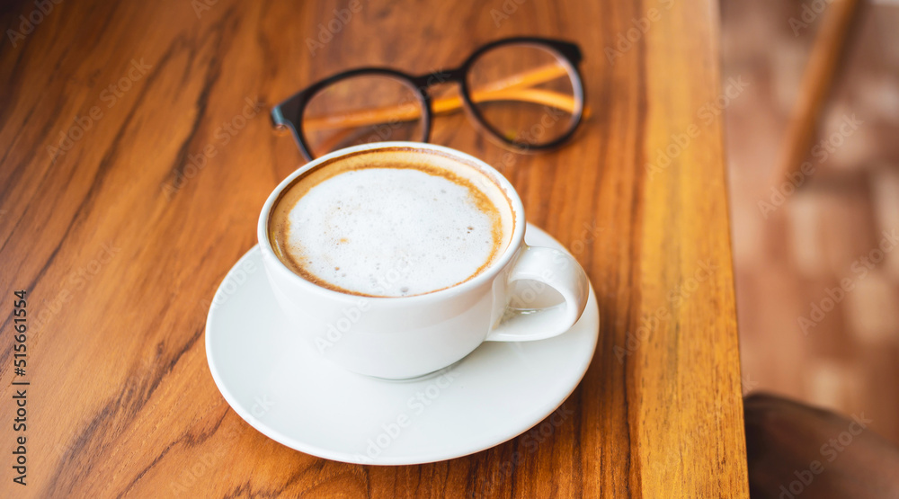 Obraz premium Cup of cappuccino coffee with latte and glasses of a woman on a wooden bar counter in a sunlit cafe in the morning.